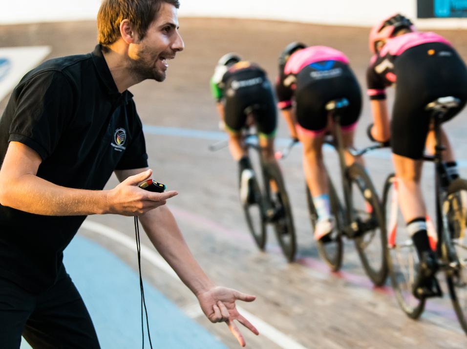 Trainer gibt Anweisungen an Gruppe von Radfahrerinnen auf einer Bahn. Vier Fahrerinnen in schwarzen und pinken Trikots.