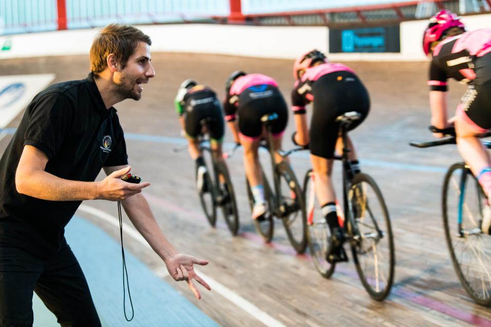 Trainer gibt Anweisungen an Gruppe von Radfahrerinnen auf einer Bahn. Vier Fahrerinnen in schwarzen und pinken Trikots.