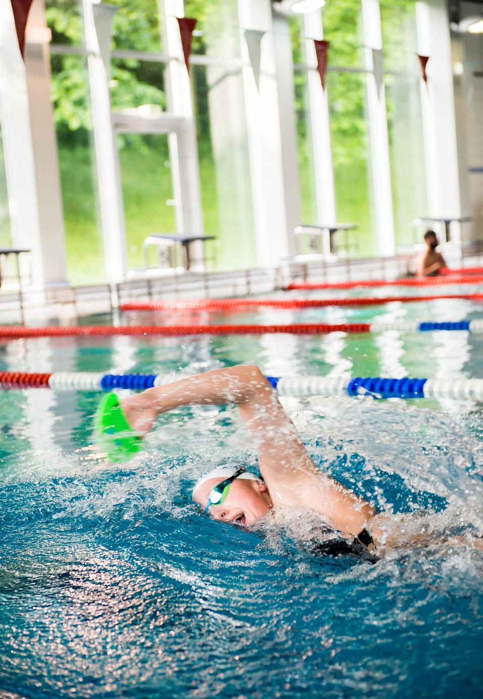 Schwimmerin mit grüner Paddel-Handschuh im Wasser eines Hallenbads, umgeben von bunten Bahnen und Fenstern.